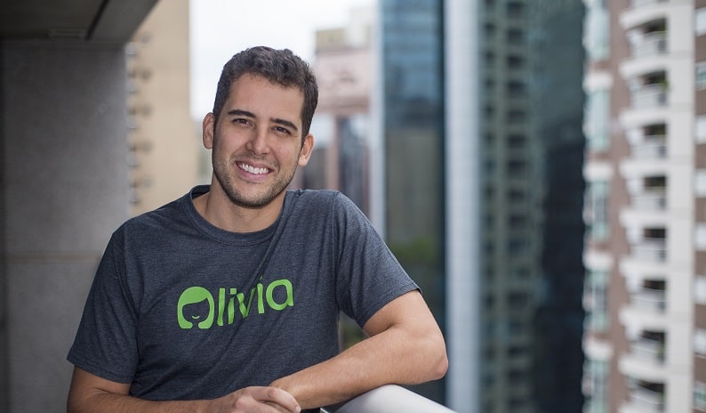 Na imagem, você vê o fundador e CEO da Olivia, Lucas Moraes. O empreendedor está posando para foto com a roupa temática da startup e, ao fundo, há diversos prédios.