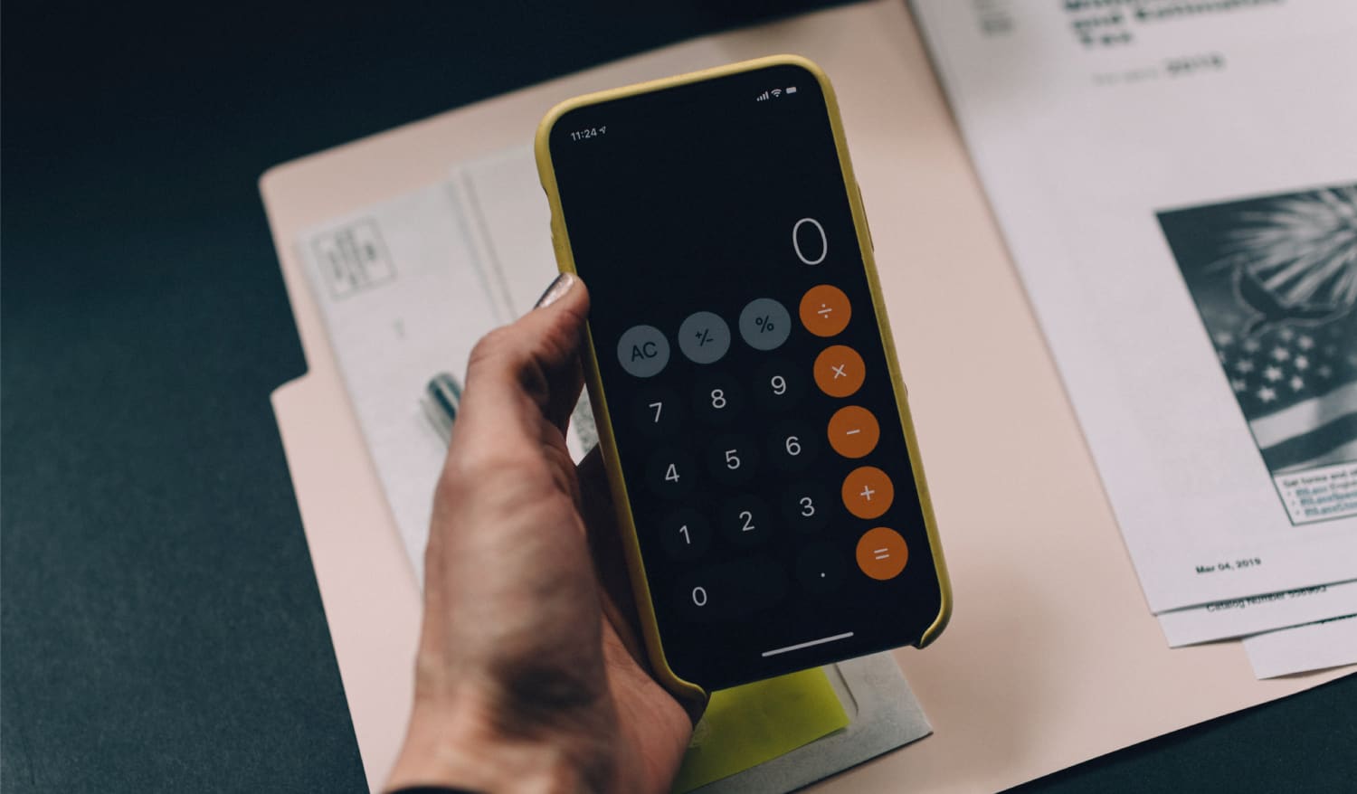 A imagem mostra uma mão segurando um celular com aplicativo de calculadora aberto. A mão está apoiada em uma mesa com outros papéis.