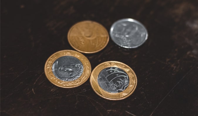 Moedas de um real, 25 centavos e 50 centavos estão em cima de uma mesa de madeira. O foco é nas moedas.