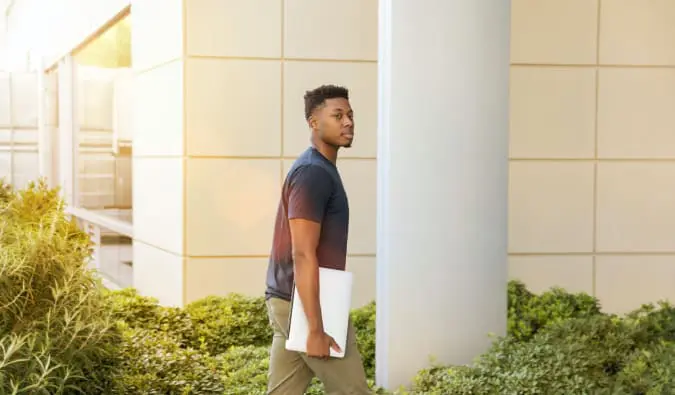 Na foto, um jovem negro está andando segurando um notebook e olha para a câmera
