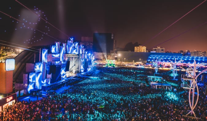 A imagem mostra um palco o Rock in Rio iluminado e uma grande quantidade pessoas assistindo no gramado