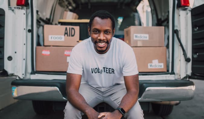 Um homem negro está sentado na parte de traz de uma van com caixas de doações. Ele usa uma camiseta escrito: volunteer, voluntário em inglês