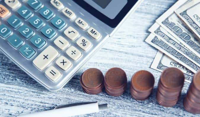 A imagem mostra uma calculadora, pilhas de moedas de dólar e notas de dólar sobre uma mesa