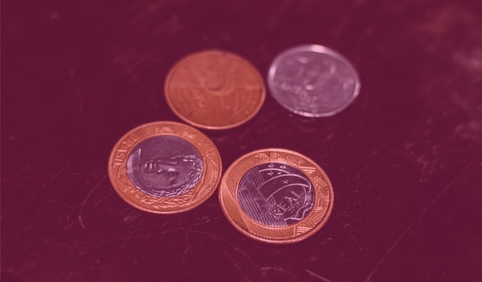 Uma foto com duas moedas de 1 real, uma moeda de 25 centavos e outra de 50 centavos. As quatro moedas estão em cima de uma superfície escura
